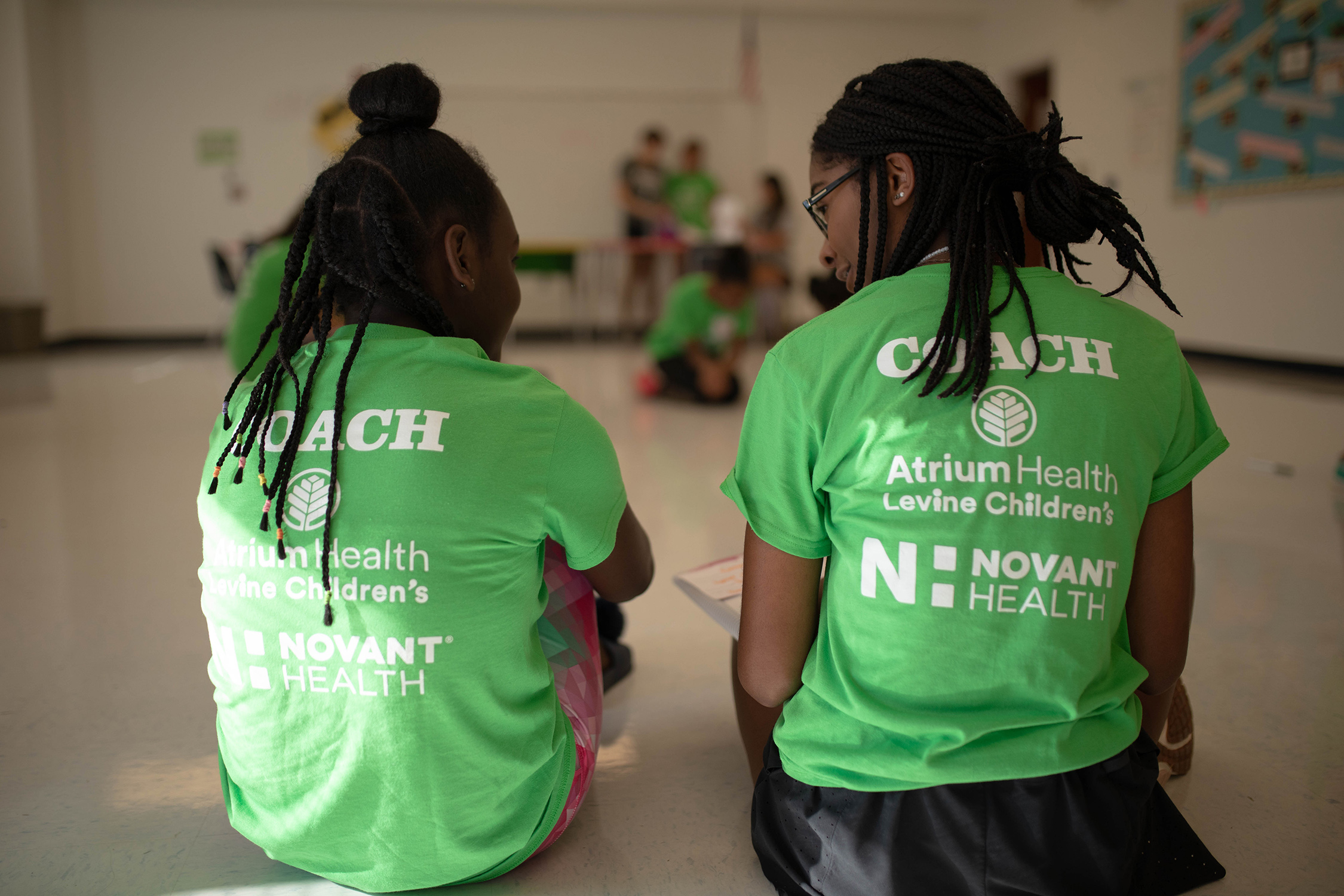 Two Girls on the Run junior coaches sit with their backs to the camera in green shirts talking to each other